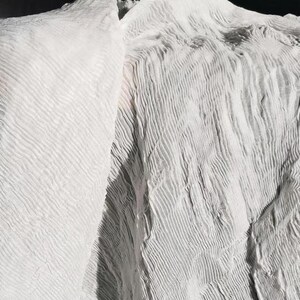 May include: Close-up of white fabric with a textured, rippled surface. The fabric appears to be draped, with folds and shadows creating depth. The material has a soft, flowing appearance.