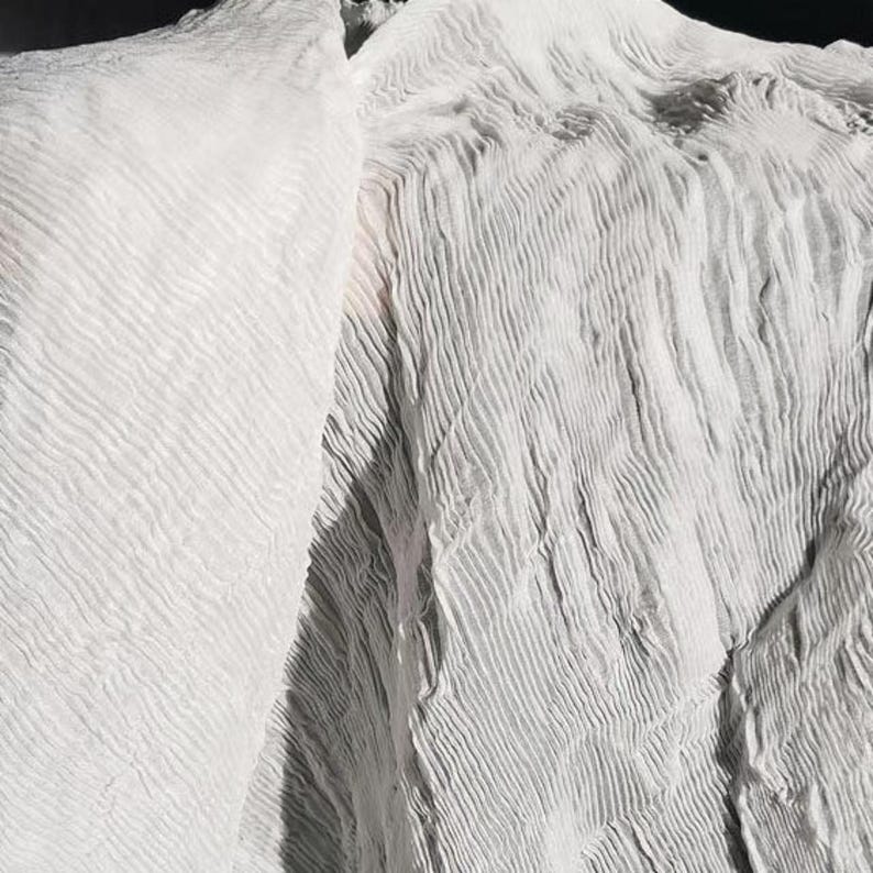 May include: Close-up of white fabric with a textured, rippled surface. The fabric appears to be draped, with folds and shadows creating depth. The material has a soft, flowing appearance.