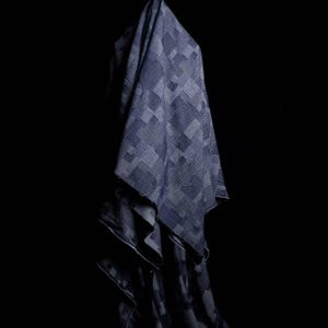 May include: A piece of fabric featuring a dark blue and grey geometric patchwork pattern. The textile displays various square and rectangular blocks with differing grid and line textures, creating a layered, draped appearance against a black background.