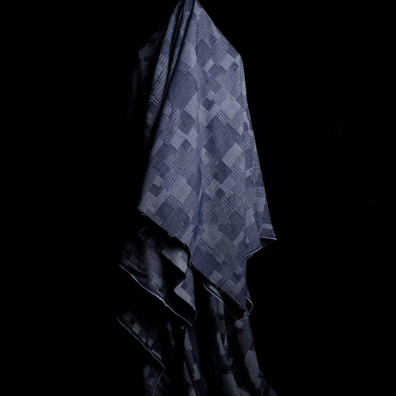 May include: A piece of fabric featuring a dark blue and grey geometric patchwork pattern. The textile displays various square and rectangular blocks with differing grid and line textures, creating a layered, draped appearance against a black background.