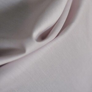 May include: Close-up of a pale pink fabric with soft folds and subtle texture. The light catches the curves, creating highlights and shadows. The material appears smooth and possibly silky, suggesting a delicate textile.