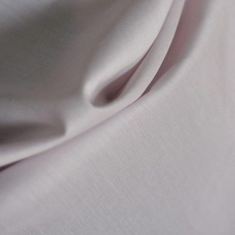 May include: Close-up of a pale pink fabric with soft folds and subtle texture. The light catches the curves, creating highlights and shadows. The material appears smooth and possibly silky, suggesting a delicate textile.