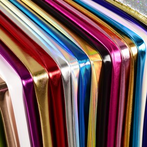 Può includere: Primo piano di una collezione di tessuti lucidi e colorati. I tessuti sono disposti in un motivo diagonale, mostrando una varietà di colori tra cui oro, rosso, argento, blu e viola. I materiali hanno un aspetto riflettente e metallico.