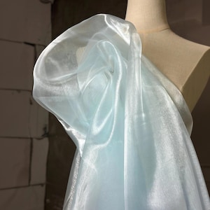 Può includere: Tessuto azzurro trasparente drappeggiato su un manichino. Il tessuto ha una lucentezza sottile e appare leggero, con pieghe morbide e un aspetto fluido. Il tessuto è di un blu pallido, quasi pastello.