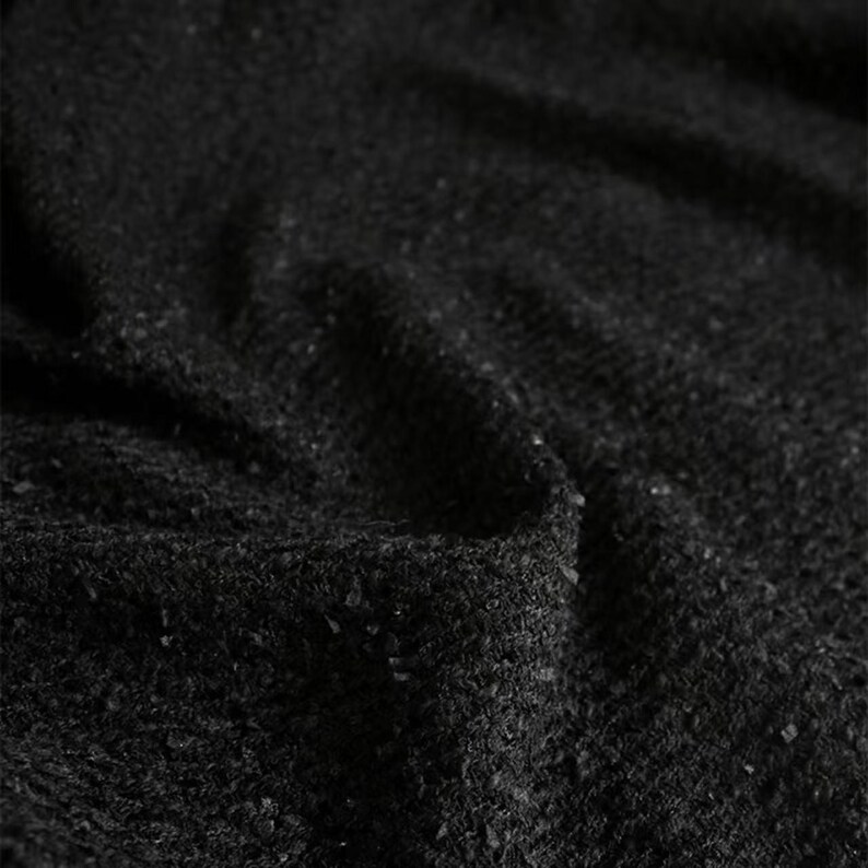 May include: Close-up of a textured black fabric with a slightly uneven surface. The fabric has a subtle sheen and a dense, woven appearance. The dark color and texture suggest a material suitable for clothing or upholstery.
