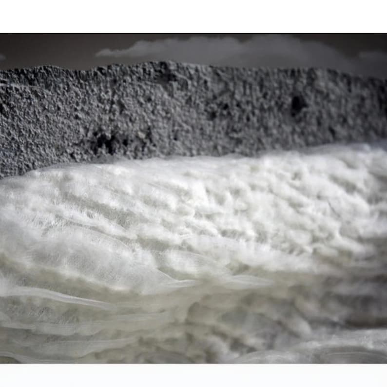 Op de afbeelding: Close-up van een doorsnede met lagen materialen. De bovenste laag is een ruw, grijs, getextureerd materiaal. Daaronder bevindt zich een dikke laag zacht, wit, pluizig materiaal. De achtergrond is een vage, neutrale tint.