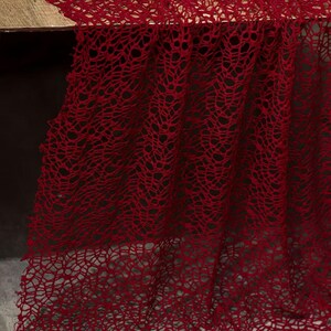 May include: A close-up of a deep red, open-weave fabric, possibly a lace or netting. The fabric is draped over a surface, with the intricate pattern and texture clearly visible. The color is a rich, saturated red.