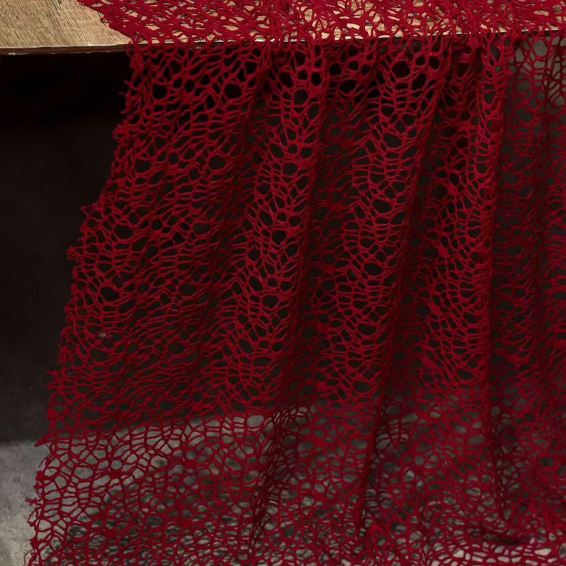 May include: A close-up of a deep red, open-weave fabric, possibly a lace or netting. The fabric is draped over a surface, with the intricate pattern and texture clearly visible. The color is a rich, saturated red.