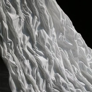 Op de afbeelding: Close-up van witte stof met getextureerde, verticale, touwachtige details. De stof is gedrapeerd en gevouwen, waardoor schaduwen en highlights ontstaan. Het algehele effect is een zachte, vloeiende textuur.