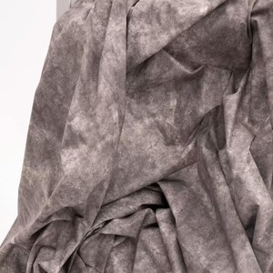 Könnte beinhalten: Nahaufnahme eines grauen Stoffes mit einem gesprenkelten Muster. Der Stoff ist drapiert und gefaltet, wodurch Schatten und Textur entstehen. Die Farbe ist ein gedämpftes Grau mit helleren und dunkleren Bereichen.