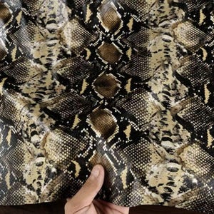 May include: Close-up of a snakeskin pattern in shades of brown, tan, and black. The texture is detailed, with scales and variations in color. The material appears to be leather or a similar fabric, with a naturalistic design.