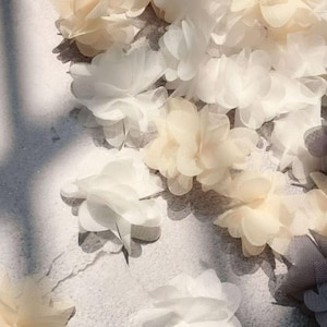 Puede incluir: Primer plano de delicadas flores de tela en capas en tonos blanco, crema y gris claro. Las flores están agrupadas, creando una superficie suave y texturizada. Ideal para manualidades o adornos.