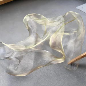 Puede incluir: Una tela fluida y transparente en tonos amarillo pálido y blanco, drapeada y dispuesta para crear curvas y pliegues suaves. El material parece delicado y ligero, con un brillo sutil. La tela está parcialmente sujeta por un palo de madera.