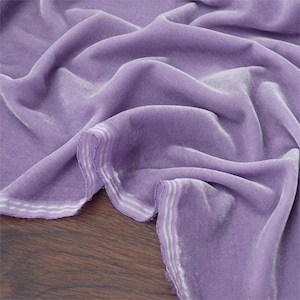 May include: A draped piece of lavender velvet fabric. The fabric has a soft, lustrous appearance with visible folds and shadows. The edges are finished with a white and light grey striped border.