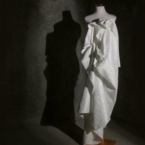 K&ouml;nnte beinhalten: Ein wei&szlig;es, drapiertes Kleidungsst&uuml;ck auf einer Schaufensterpuppe. Der Stoff hat ein subtiles Muster und f&auml;llt in weichen Falten. Die Schaufensterpuppe steht auf einem Holzst&auml;nder vor einem dunkelgrauen Hintergrund mit einem ausgepr&auml;gten Schatten.