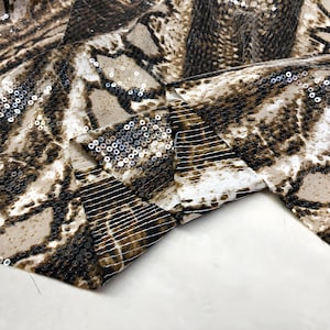 May include: A close-up shot of a fabric featuring a snakeskin design in brown, tan, and white. The material is adorned with small, reflective sequins. The fabric is folded, showcasing its texture and pattern. The overall effect is one of luxury.