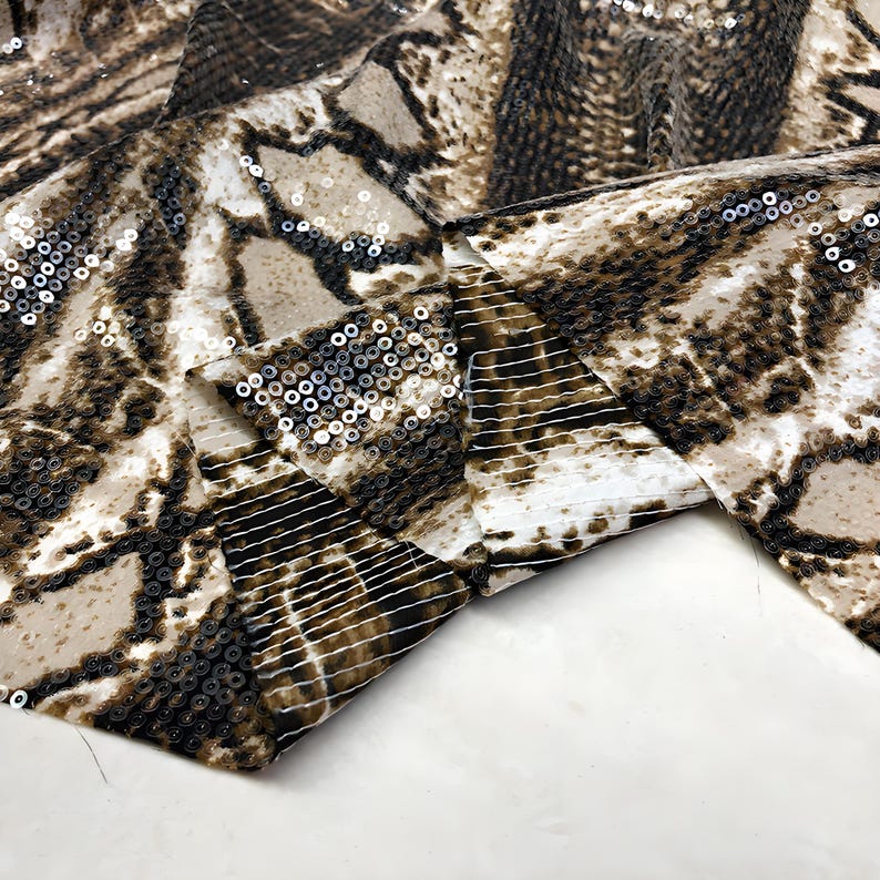May include: A close-up shot of a fabric featuring a snakeskin design in brown, tan, and white. The material is adorned with small, reflective sequins. The fabric is folded, showcasing its texture and pattern. The overall effect is one of luxury.