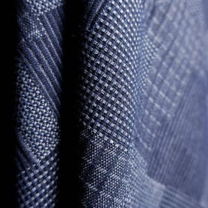 May include: A close-up view of a dark blue fabric featuring a complex woven texture. The material displays a repeating geometric pattern composed of small, raised squares and diagonal lines, creating a dense, tactile surface with deep blue and white threads.