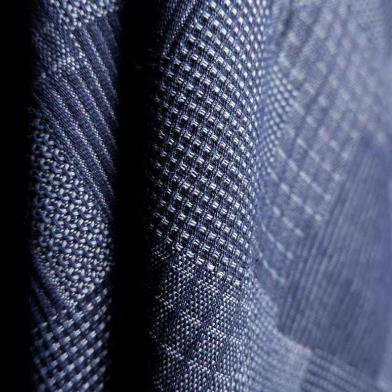 May include: A close-up view of a dark blue fabric featuring a complex woven texture. The material displays a repeating geometric pattern composed of small, raised squares and diagonal lines, creating a dense, tactile surface with deep blue and white threads.