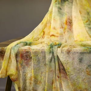 May include: A flowing, sheer fabric with a crinkled texture. The fabric features a watercolor-like pattern with yellow, green, and red hues. The material is draped over a surface, creating soft folds and shadows.