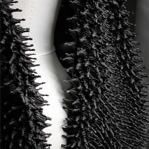 May include: A close-up of a black, textured garment. The fabric has a dense, spiky appearance, with a three-dimensional quality. The garment appears to be draped over a white form, creating a contrast between the dark texture and the smooth, light background.