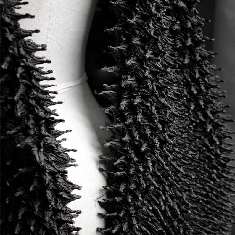 May include: A close-up of a black, textured garment. The fabric has a dense, spiky appearance, with a three-dimensional quality. The garment appears to be draped over a white form, creating a contrast between the dark texture and the smooth, light background.