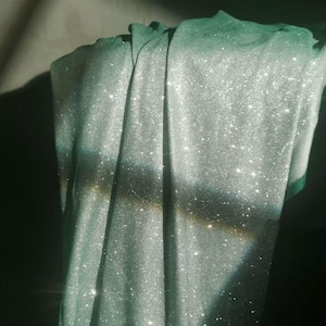 Op de afbeelding: Een gedrapeerde stof met een glinsterende, glitterende textuur. Het materiaal is een zachte, lichtgroene kleur, met een subtiele glans. De stof is gevouwen, waardoor schaduwen en highlights ontstaan die de textuur benadrukken. Het totale effect is elegant en luxueus.