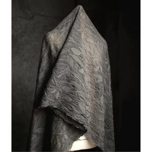 May include: A draped, gray fabric with a leaf pattern. The fabric is textured and appears to be made of a soft material. The edges of the fabric are unfinished, giving it a raw, natural look. The fabric is draped over a form, creating folds and shadows.