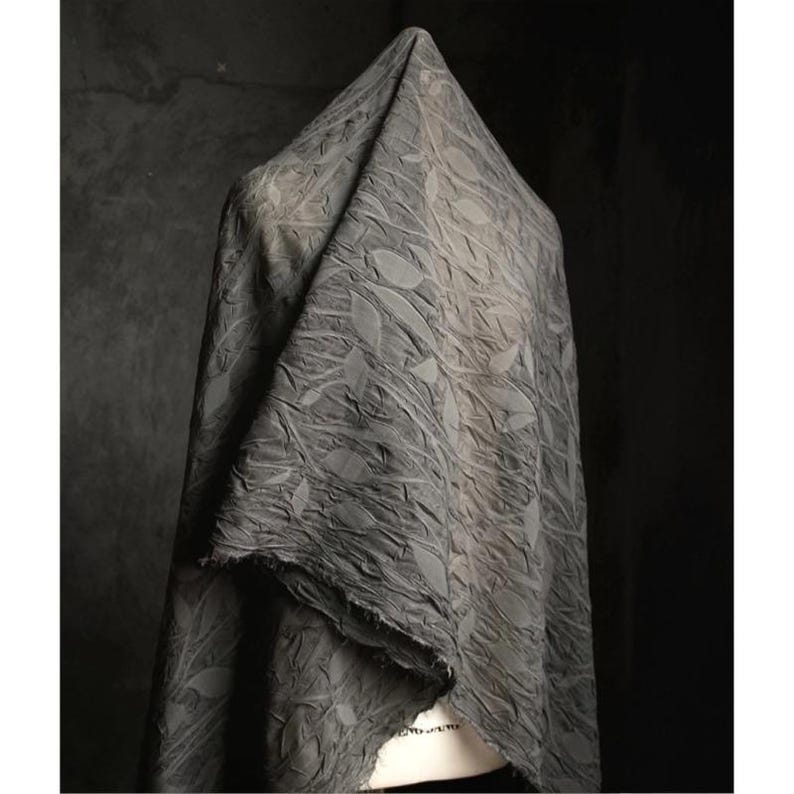May include: A draped, gray fabric with a leaf pattern. The fabric is textured and appears to be made of a soft material. The edges of the fabric are unfinished, giving it a raw, natural look. The fabric is draped over a form, creating folds and shadows.