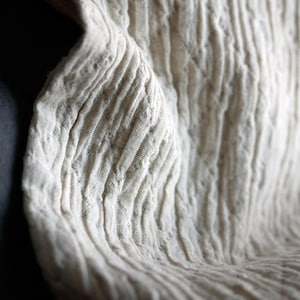 May include: Close-up of a textured, off-white fabric with vertical, ribbed patterns. The fabric appears soft and slightly crumpled, with subtle variations in tone and texture. The overall effect is of a natural, organic material.