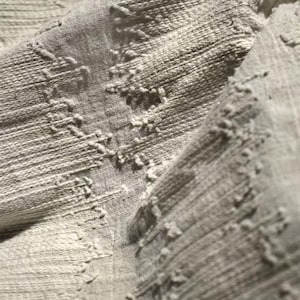 May include: Close-up of a textured, light gray fabric. The fabric has a woven appearance with raised, irregular patterns. The texture is visible, with a mix of horizontal lines and raised, textured elements. The overall look is soft and neutral.