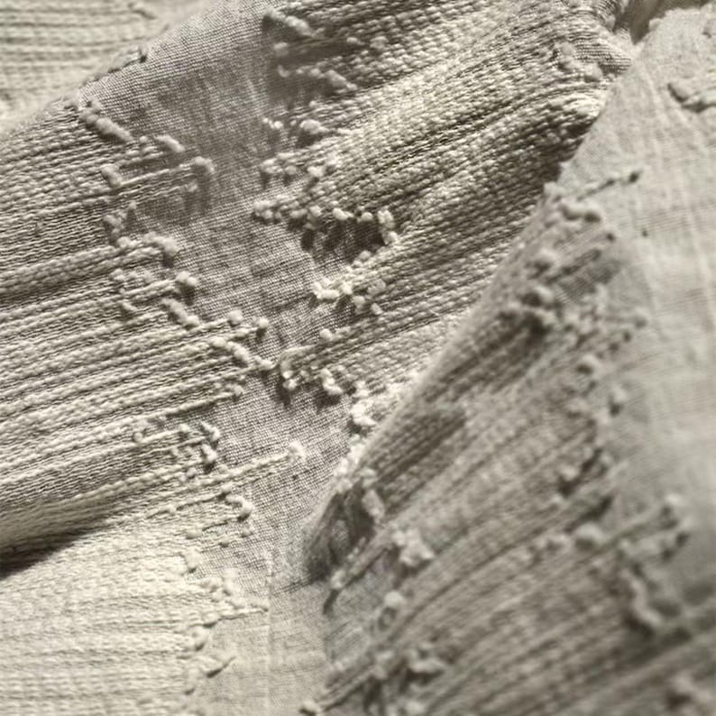May include: Close-up of a textured, light gray fabric. The fabric has a woven appearance with raised, irregular patterns. The texture is visible, with a mix of horizontal lines and raised, textured elements. The overall look is soft and neutral.