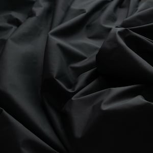 May include: Close-up of a black fabric with soft folds and shadows. The fabric appears to be a solid color, with a smooth texture. The image highlights the fabric's drape and the way light interacts with it.