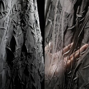 May include: Close-up of a dark gray fabric with a textured, crinkled surface. The fabric appears to be draped, with folds and ridges creating a dynamic pattern. The texture is rough and uneven, with a matte finish. A hand is visible through the sheer fabric.