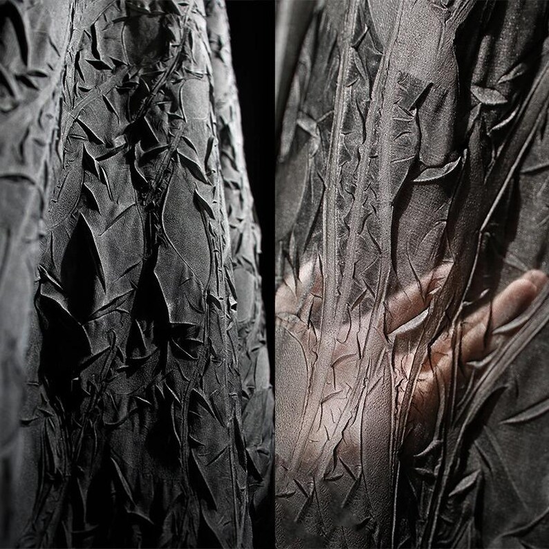 May include: Close-up of a dark gray fabric with a textured, crinkled surface. The fabric appears to be draped, with folds and ridges creating a dynamic pattern. The texture is rough and uneven, with a matte finish. A hand is visible through the sheer fabric.