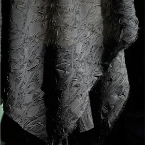 May include: A close-up shot of a dark gray fabric with a textured, raised pattern. The fabric appears to be draped, with visible folds and shadows. The edges of the fabric have a raw, unfinished look, adding to its unique texture.