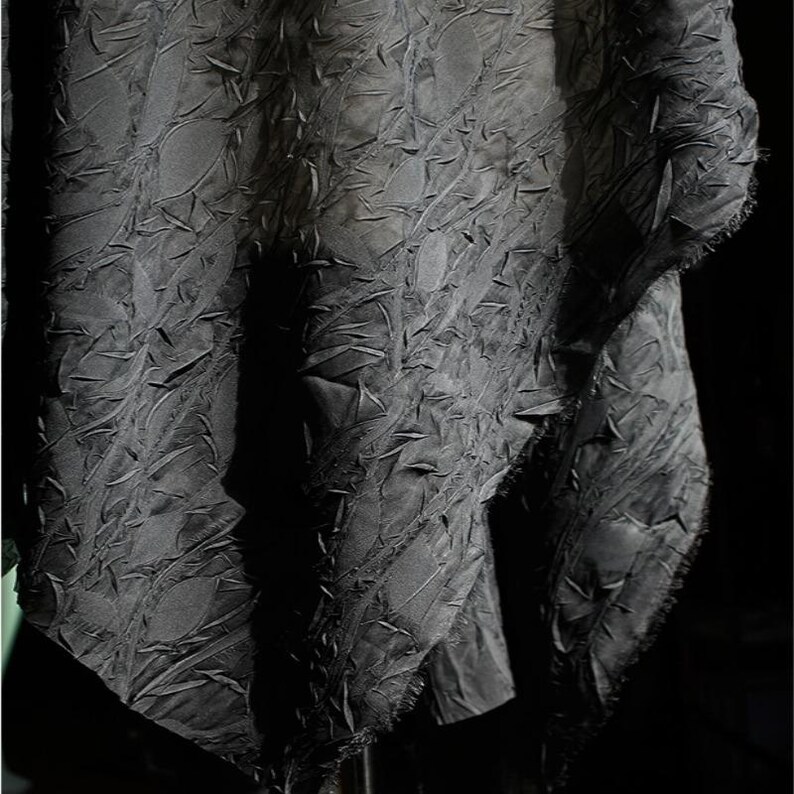 May include: A close-up shot of a dark gray fabric with a textured, raised pattern. The fabric appears to be draped, with visible folds and shadows. The edges of the fabric have a raw, unfinished look, adding to its unique texture.