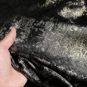 Puede incluir: Primer plano de tela negra con un patr&oacute;n dorado brillante y moteado. La tela es suave y parece estar drapeada, con pliegues y texturas visibles. El efecto general es lujoso y elegante.