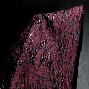 May include: Close-up of a textured fabric in a deep burgundy color with a glossy, almost wet appearance. The fabric has a raised, irregular pattern with hints of dark gray or black woven throughout. The overall effect is rich and luxurious.