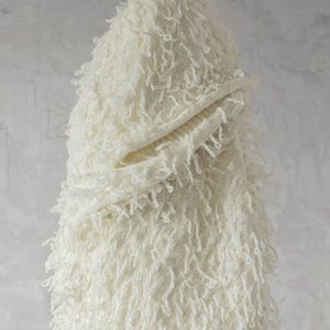 May include: Off-white knitted hooded scarf with a textured, fringed design. The scarf has a hood and a wrap-around design, offering warmth and style. The soft, cream color and unique texture make it a cosy accessory for cooler weather.