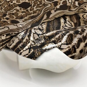 May include: A close-up of a fabric with a snakeskin pattern in shades of brown, tan, and black. The fabric is draped over a white, curved form, creating folds and shadows. The fabric has a shiny, sequined texture.