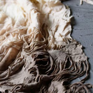 Puede incluir: Primer plano de una tela texturizada en tonos crema y marrón. La tela tiene un borde deshilachado y con flecos y una apariencia en capas, creando un efecto suave y drapeado. El textil parece ser lino o una fibra natural similar.