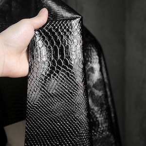 Peut inclure: Gros plan sur un morceau de similicuir noir avec un motif peau de serpent. Le matériau est maintenu, montrant la texture et l'éclat du tissu. Les écailles sont clairement définies et le matériau semble souple.