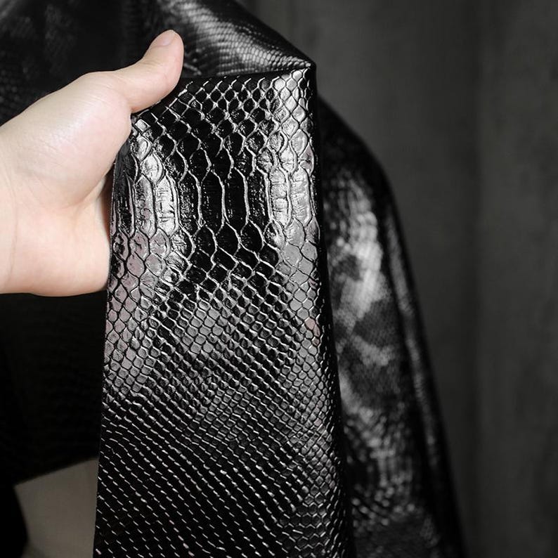 Może przedstawiać: Zbliżenie kawałka czarnej sztucznej sk&oacute;ry ze wzorem sk&oacute;ry węża. Materiał jest trzymany, pokazując fakturę i połysk tkaniny. Łuski są wyraźnie zdefiniowane, a materiał wydaje się elastyczny.