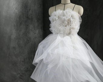 May include: A white, layered tulle gown on a mannequin. The bodice features a large, ruffled floral design. The skirt is tiered, creating a flowing silhouette. The dress is displayed against a dark gray backdrop, highlighting the delicate fabric.