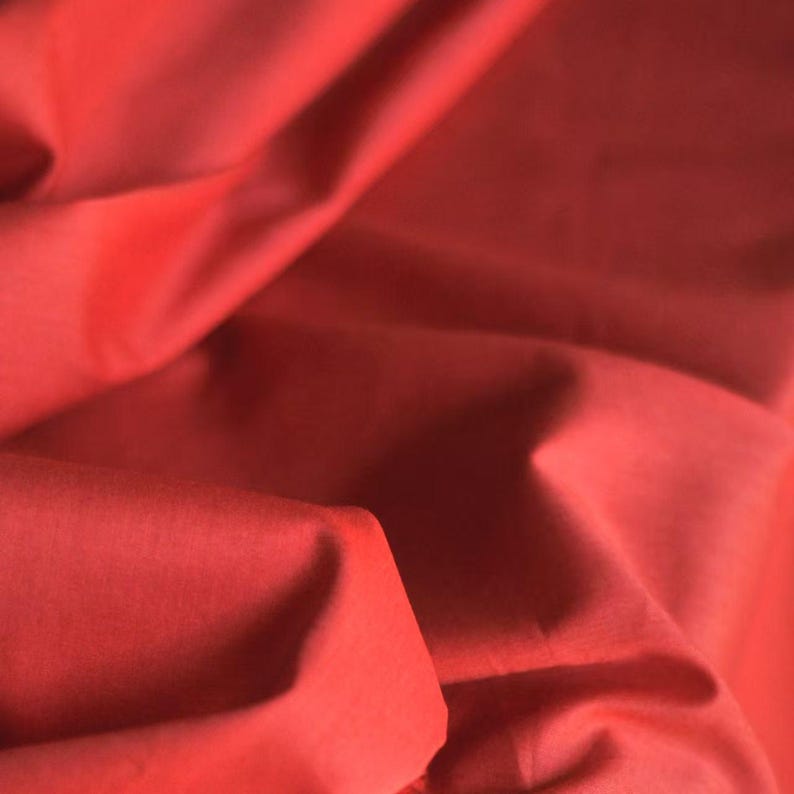 May include: Close-up of a vibrant red fabric with soft folds and shadows. The material appears smooth and possibly silky, with a rich, saturated color. The fabric is likely used for clothing or home decor.