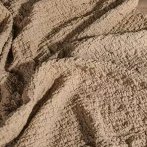 Puede incluir: Primer plano de una tela beige texturizada con aspecto arrugado. La tela tiene un patrón tejido y está drapeada, creando sombras y reflejos. El material parece suave y podría usarse para la decoración del hogar.