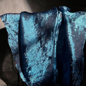 Op de afbeelding: Een gedrapeerde teal stof met een getextureerde, gekreukelde uitstraling. De stof heeft een glinsterende, iriserende kwaliteit, met highlights en schaduwen die diepte creëren. Het materiaal lijkt gevouwen en vloeiend, wat duidt op een decoratief textiel.