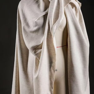 May include: A light beige fabric is draped over a dress form, creating soft folds and a flowing appearance. The fabric has a textured finish. The dress form is a neutral colour, and the background is dark.