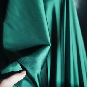 Puede incluir: Primer plano de tela verde esmeralda con una textura suave y brillante. La tela está doblada, creando sombras y reflejos que enfatizan su caída y brillo. La imagen muestra el color rico y la calidad de la tela.